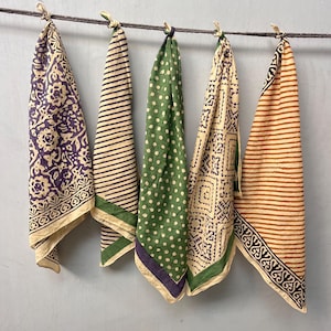 Puede incluir: Cinco pañuelos estampados colgados de una cuerda. Los pañuelos presentan varios diseños, incluyendo rayas, lunares y estampados florales, en colores como morado, verde y beige. Cada pañuelo está anudado en la parte superior.