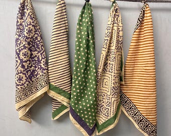 May include: Five patterned bandanas hanging from a rope. The bandanas feature various designs, including stripes, polka dots, and floral patterns, in colors like purple, green, and tan. Each bandana is knotted at the top.