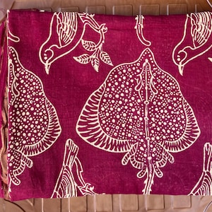 May include: Folded burgundy fabric with a repeating white pattern of stylized birds and leaf-like shapes. The fabric has a fringed edge and is neatly stacked, showcasing the detailed design.