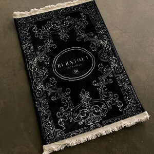 May include: Black and white rug with a fringed border. The rug features a traditional ornate design with a circular logo in the center that reads "BURNOUT ISTANBUL" with a stylized "B" inside the circle.