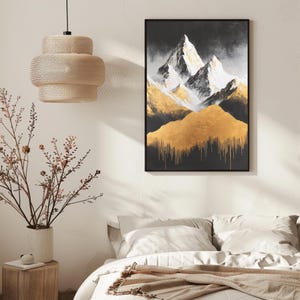 Op de afbeelding: Een ingelijst abstract bergschilderij met gouden en witte accenten. Het kunstwerk heeft een zwarte achtergrond met goud dat van de bergen afdruipt. Het schilderij wordt tentoongesteld boven een bed met witte lakens.