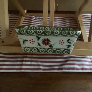May include: A white ceramic baking dish with a green and red floral pattern. The dish is sitting on a red and white striped fabric.