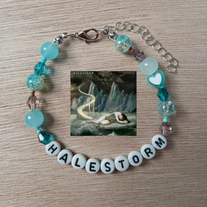 May include: A beaded bracelet featuring a square image of the Halestorm album cover, light blue and teal glass beads, clear faceted beads, a white heart bead, and white letter beads spelling HALESTORM with a silver metal lobster clasp.