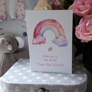 May include: A white card with a watercolour rainbow, clouds, and silver glitter accents. The text reads "Welcome to the World Poppy Mae 23.08.23".