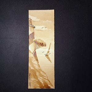 Puede incluir: Marcapáginas de acuarela en tonos sepia con un faro en un acantilado. La obra de arte incluye un árbol, pájaros y la firma del artista, "RANDELL 2023". El marcapáginas es rectangular y tiene un borde texturizado.