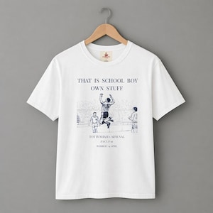 Puede incluir: Camiseta blanca con cuello redondo y mangas cortas. La camiseta presenta una ilustración azul marino de un jugador de fútbol en acción, con texto encima y debajo de la imagen. El texto dice "THAT IS SCHOOL BOY OWN STUFF" y "TOTTENHAM v ARSENAL FA CUP 61 WEMBLEY 14 APRIL".