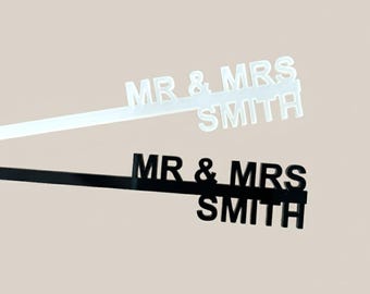 May include: Two wedding cake toppers, one white and one black, each with the text "MR & MRS SMITH". The toppers are on thin, rectangular sticks, ideal for inserting into a cake. The background is a solid, neutral color.