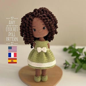 May include: A handmade crochet doll with brown skin, brown curly hair, and large eyes. The doll wears a green dress with a white bow and trim. The text "AMY CROCHET DOLL PATTERN" is visible. The doll is on a wooden heart-shaped base.