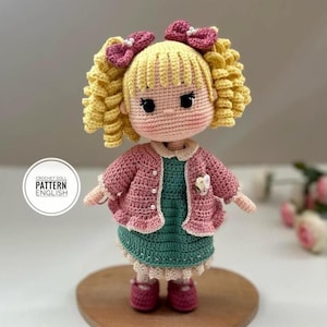 Pode incluir: Boneca de crochê feita à mão com cabelo loiro encaracolado, laços rosa e um cardigan rosa sobre um vestido verde. A boneca tem olhos pretos de botão. O texto "PATTERN ENGLISH" está na boneca.