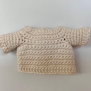 Pu&ograve; includere: Un piccolo maglione all'uncinetto color crema con maniche corte.