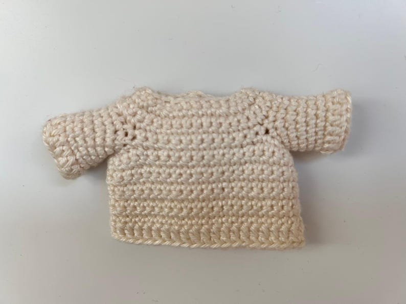 Pu&ograve; includere: Un piccolo maglione all'uncinetto color crema con maniche corte.