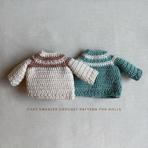 May include: Two miniature crochet sweaters, one cream with brown stripes and the other teal with white stripes. The text "COZY SWEATER CROCHET PATTERN FOR DOLLS" is displayed below the sweaters. These doll sweaters are handmade.