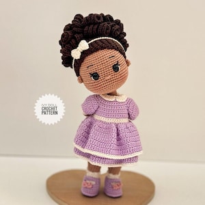 Może przedstawiać: Ręcznie robiona szydełkowa lalka z brązową skórą, ciemnymi kręconymi włosami i białą opaską z kokardą. Lalka ma na sobie lawendową sukienkę z białymi wykończeniami i pasujące buty. Widoczny jest tekst "IVY DOLL CROCHET PATTERN".