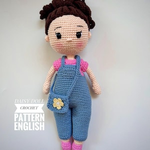 May include: Crocheted doll with brown hair, wearing a pink shirt, blue dungarees, and a yellow flower purse. The doll is standing on a white background. Daisy Doll Crochet Pattern English.