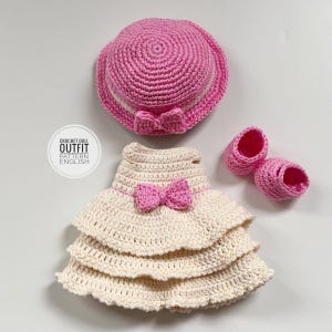May include: A handmade crochet doll outfit in cream and pink. The set includes a tiered dress with a pink bow, a matching hat with a bow, and a pair of small shoes. The text "CROCHET DOLL OUTFIT PATTERN ENGLISH" is visible.