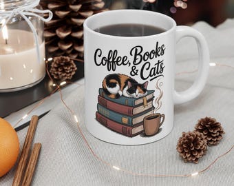 May include: White ceramic mug with the words "Coffee, Books & Cats" and a graphic of a cat sleeping on a stack of books. A small brown coffee cup is next to the books. The mug is on a light gray surface with decorative items.
