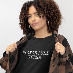 May include: A person wearing a black t-shirt with the text "BACKGROUND EXTRA" in white lettering. They are also wearing a brown and black checked shirt.