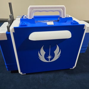 May include: A blue plastic storage case with a white handle and a white Jedi symbol on the front. The case has a white trim and a white latch.