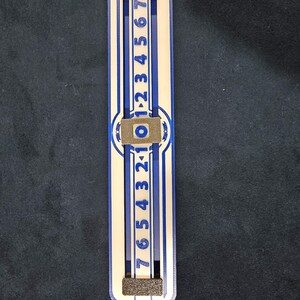 Puede incluir: Un deslizador de números de madera blanco y azul con los números del 0 al 7 impresos en él. El deslizador está hecho de madera y tiene un diseño a rayas azul y blanco. El deslizador se utiliza para contar números.