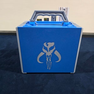 May include: A blue carrying case with a silver Mandalorian symbol on the front. The case has a grey handle and a silver trim.