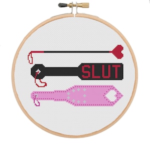 May include: A cross-stitch pattern on a wooden embroidery hoop with two paddles. The top paddle is black with the word "SLUT" in red letters. The bottom paddle is pink with a heart shape at the end.
