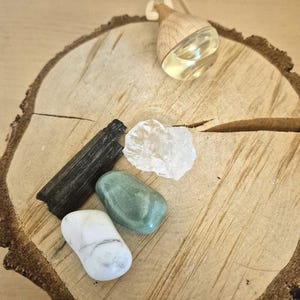 Könnte beinhalten: Ein ätherisches Öl Diffusor mit einem Holzaufsatz und einem Glasbehälter, der mit einer gelben Flüssigkeit gefüllt ist. Der Diffusor liegt auf einer Holzscheibe neben verschiedenen Kristallen und Steinen, darunter ein schwarzer Turmalin, ein klarer Quarz und grüne und weiße Trommelsteine.
