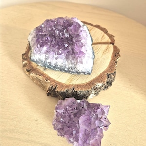 Könnte beinhalten: Zwei Amethyst-Kristallcluster. Einer ist eine große, halbkreisförmige Geode auf einer Holzscheibe, die tiefviolette Kristallspitzen zeigt. Der andere ist ein kleinerer, freiformiger Cluster der gleichen Farbe. Beide befinden sich auf einer hellen Holzoberfläche.