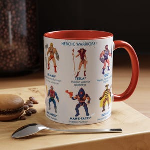 Può includere: Tazza in ceramica bianca con interno e manico rossi, con illustrazioni di He-Man e altri personaggi della serie "Heroic Warriors". La tazza è su una superficie di legno con un cucchiaio e un pasticcino.