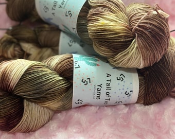 May include: Close-up of two skeins of yarn with a gradient of brown, tan, and cream colors. Each skein has a label that reads "A Tail of Two Yarns". The yarn is resting on a pink, textured surface.