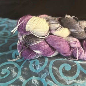 May include: A skein of hand-dyed yarn in shades of purple, cream, and gray. The yarn is arranged in a loose bundle, with the colors blending together. The yarn is sitting on a teal fabric with a swirling pattern.