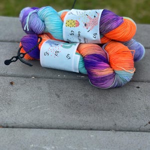May include: Two skeins of colourful yarn in shades of purple, orange, blue, and teal. Each skein has a label with a cactus and llama design. The yarn is displayed on a grey surface.