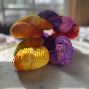 May include: Three skeins of yarn in shades of yellow, orange, purple, and pink. The yarn is arranged in a pile, with the yellow skein on top.
