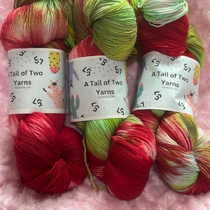 Puede incluir: Tres ovillos de hilo en combinaciones de colores rojo, verde y blanco. El hilo está teñido a mano y tiene un aspecto moteado. Cada ovillo está etiquetado con "A Tail of Two Yarns" con la ubicación de la empresa, Valdosta, GA.