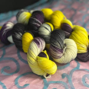 以下が含まれることがあります： 黄色、紫、黒、クリーム色の手染め糸の束。 糸は、青い渦模様のピンク色の布の上に配置されています。 糸は柔らかくふわふわで、編み物や編み物のプロジェクトに最適です。