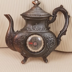 May include: A silver teapot-shaped thermometer with a round dial and a black needle. The thermometer has a decorative design with floral accents and a black finish.