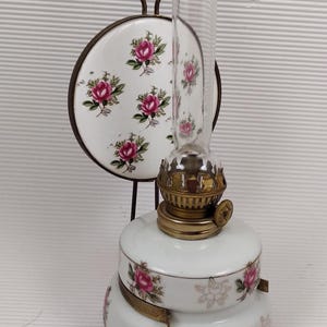 May include: A vintage oil lamp with a clear glass chimney and a white ceramic base. The base and a matching decorative plate feature pink rose floral patterns. The lamp has brass accents and a metal handle.