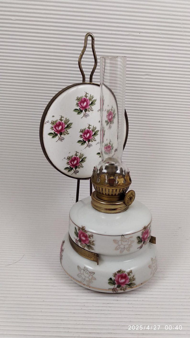 May include: A vintage oil lamp with a clear glass chimney and a white ceramic base. The base and a matching decorative plate feature pink rose floral patterns. The lamp has brass accents and a metal handle.