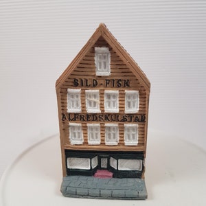 Könnte beinhalten: Ein kleines, braunes Holzgebäude mit weißen Fenstern und einem Schild mit der Aufschrift "SILD-FISK ALFREDSKULSTAD". Das Gebäude hat einen grauen Steinsockel und eine rote Tür.