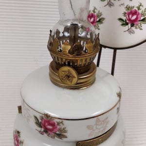 May include: A vintage oil lamp with a white porcelain base and a clear glass chimney. The lamp is decorated with pink roses and gold accents. The lamp has a brass burner and a decorative metal stand.