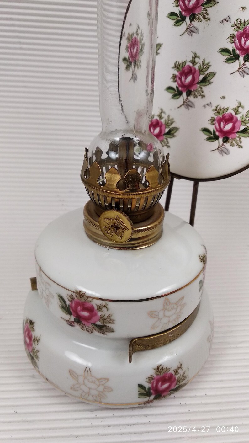 May include: A vintage oil lamp with a white porcelain base and a clear glass chimney. The lamp is decorated with pink roses and gold accents. The lamp has a brass burner and a decorative metal stand.