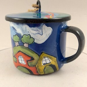 Puede incluir: Una taza de esmalte azul con tapa que presenta una escena pintada a mano de casas coloridas, colinas verdes y árboles bajo un cielo azul con nubes blancas. La taza tiene un asa negra y un borde negro, con un trozo de cuerda en la tapa.