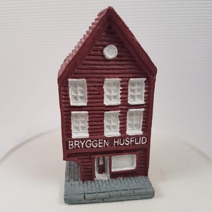 Könnte beinhalten: Ein rotes Holzhaus im Miniaturformat mit weißen Verzierungen und einem Schild mit der Aufschrift "Bryggen Husflid". Das Haus hat einen weißen Kreis auf der Vorderseite und eine kleine Tür.