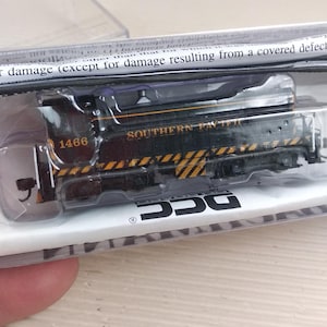 May include: A black model train engine with yellow and white warning stripes. The engine has the number 1466 and the words "SOUTHERN PACIFIC" printed on the side. The train is packaged in a clear plastic case with the word "DCC" printed on the bottom.