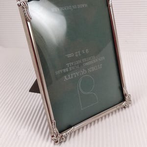 Peut inclure: Cadre photo en métal argenté avec des détails ornés. Le cadre est rectangulaire et contient un fond vert. Le texte "JYDEN QUALITY" et "MADE IN DENMARK" sont visibles. Le cadre mesure 9 x 12 cm.