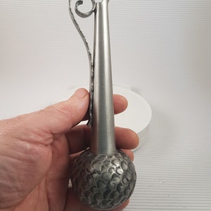 K&ouml;nnte beinhalten: Eine silberne Metallvase mit einem strukturierten, runden Boden und einem langen, schlanken Hals. Die Vase hat einen dekorativen, gekr&auml;uselten Griff.