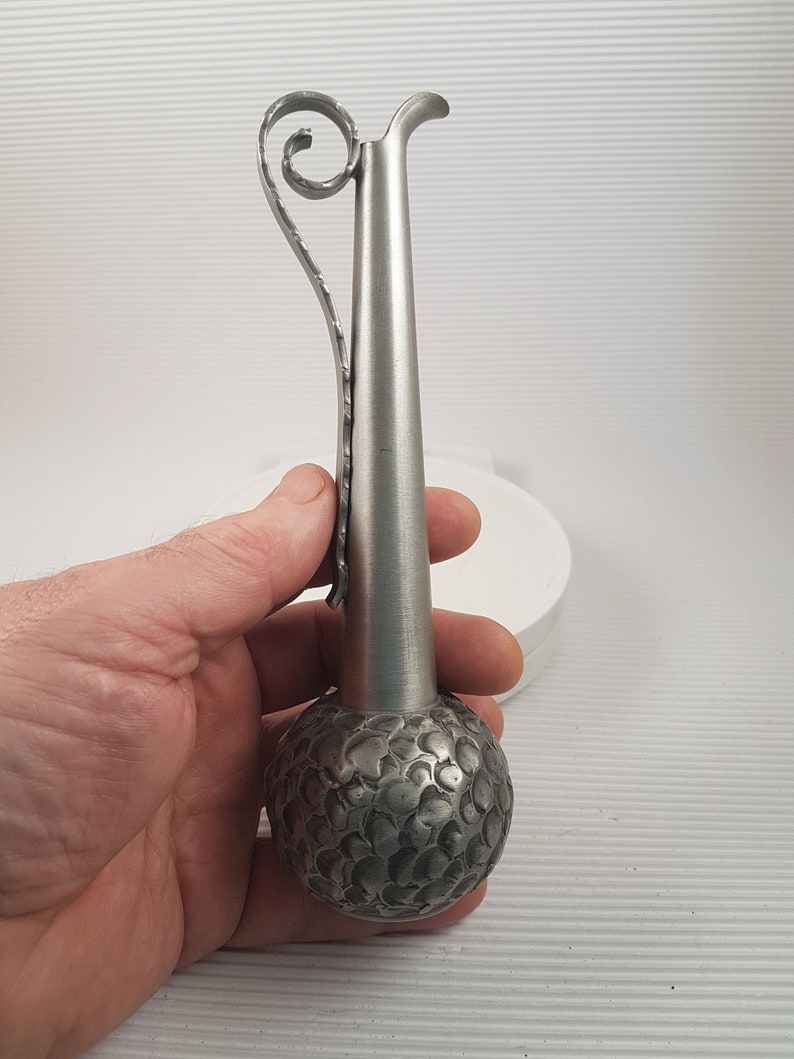 K&ouml;nnte beinhalten: Eine silberne Metallvase mit einem strukturierten, runden Boden und einem langen, schlanken Hals. Die Vase hat einen dekorativen, gekr&auml;uselten Griff.
