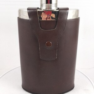 Op de afbeelding: Een bruine met leer beklede fles met een zilveren bovenkant en een bijpassende leren etui. De fles heeft een afgeronde rechthoekige vorm en een zilveren dop. De leren etui heeft een knoopsluiting. De fles is ontworpen om vloeistoffen in te bewaren.