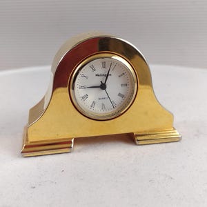 Può includere: Un orologio al quarzo color oro con quadrante bianco e numeri romani. L'orologio ha una base rettangolare e una parte superiore curva. Il marchio "Wellington" è stampato sul quadrante.