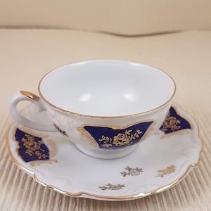 May include: A white teacup and saucer with a blue and gold floral design. The cup has a gold rim and the saucer has a scalloped edge.