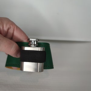 Könnte beinhalten: Eine silberne Flasche mit einem schwarzen Band und einer grünen Ledertasche.